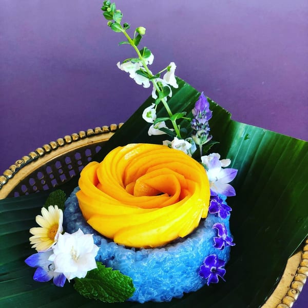 Blue sticky rice with mango slices arranged like a rose, garnished with flowers and mint on a banana leaf.