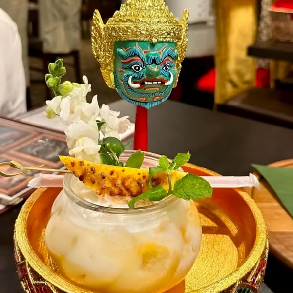 A Thai dessert with pineapple and mint, served in a glass with a decorative mask behind it.