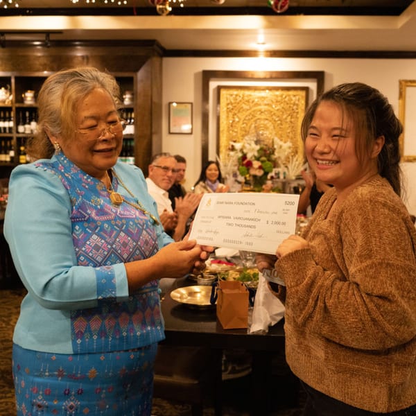 Two women smiling, one presenting a large check to the other, in a restaurant setting.