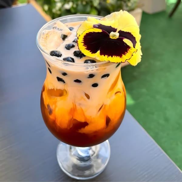 Thai iced tea with boba and a yellow pansy garnish, on a gray table outdoors.