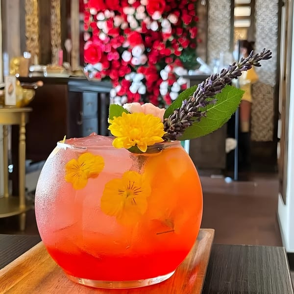 Vibrant orange and red cocktail in a round glass, garnished with flowers and lavender, on a wooden tray.