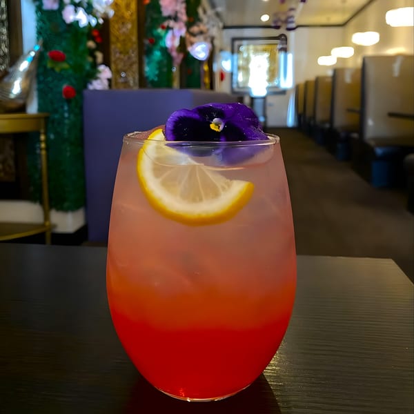 Vibrant red and pink cocktail with a lemon slice and purple flower garnish on a dark table.