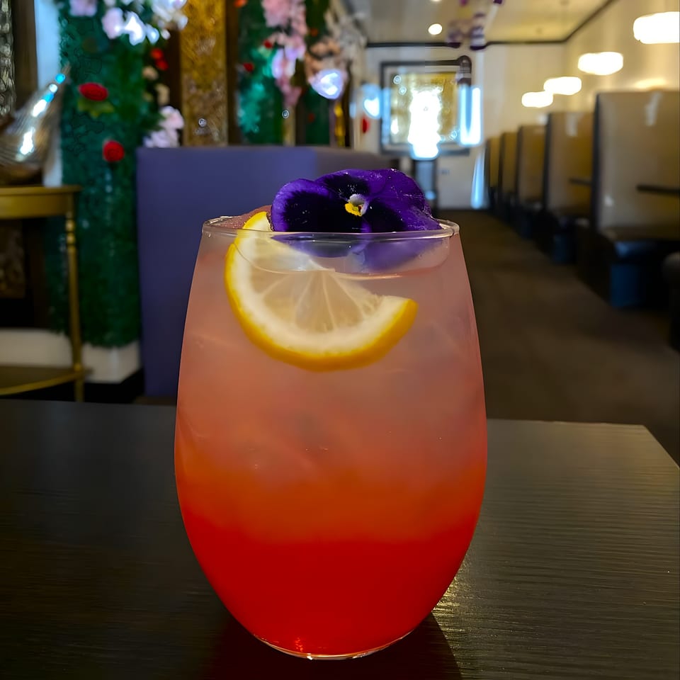 Vibrant red and pink cocktail with a lemon slice and purple flower garnish on a dark table.